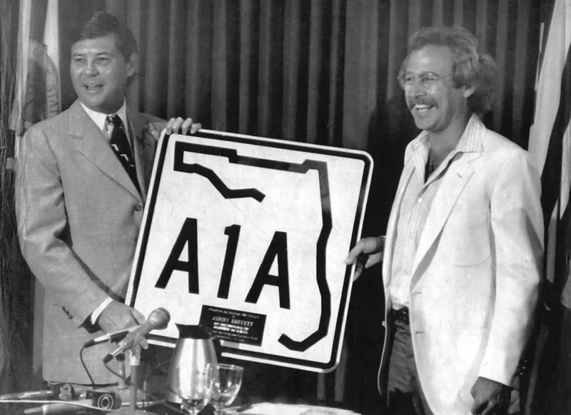 Governor Bob Graham presents musician Jimmy Buffett with a highway A1A sign after they teamed up to found "Save the Manatee Club" in 1981, leading to the creation of "Save the Manatee" license plates and their ensuing friendship.